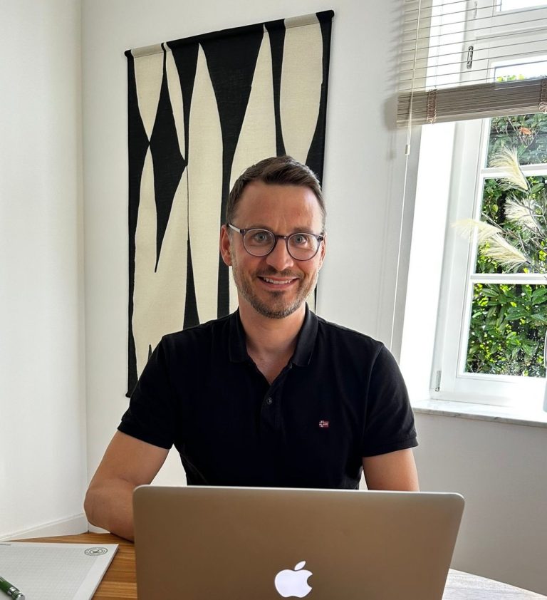Freundlich lächelnder Marc Albertz mit Brille sitzt an einem Tisch vor einem Laptop, helle Umgebung.