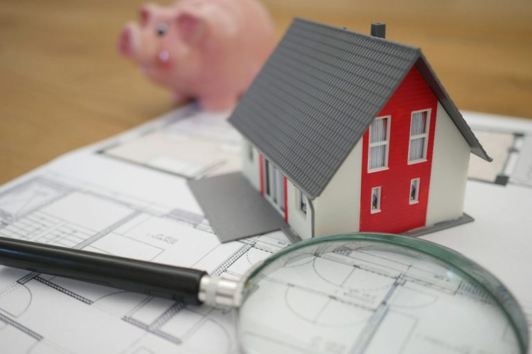 Miniaturhausmodell auf Plänen mit einer Lupe und einem Sparschwein im Hintergrund.